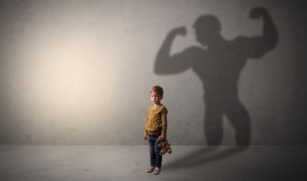 Little waggish boy in an empty room with musclemen shadow behind
