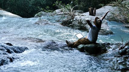 Young man using laptop computer on a waterfall and Camping. Travel and Freelance work concept.