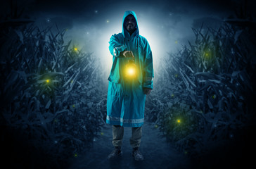 Man in raincoat at night coming from thicket and looking something with glowing lantern
