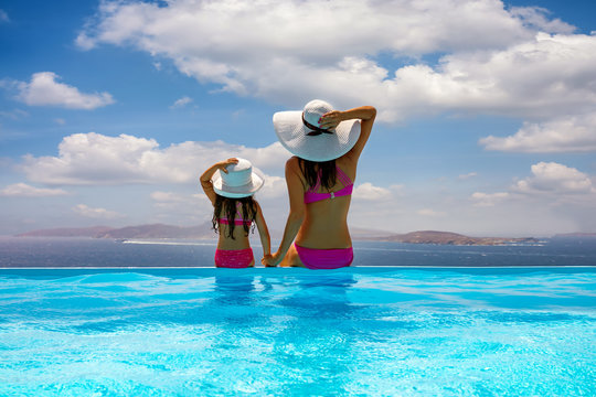 Mutter und Tochter mit Sonnenhut sitzen gemeinsam am Poolrand und genie&szlig;en die Aussicht auf das Meer