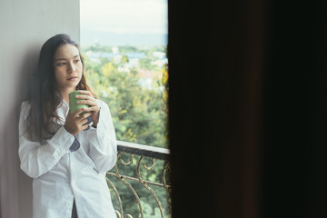 Attractive young woman holding a cup with hot tea or coffee and standing near the balcony in her home with copy space.morning coffee concept