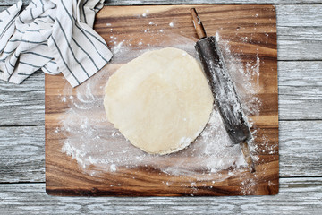 Homemade butter pie crust dough with rolling pin and towel over floured rustic wooden background. Image shot from overhead.