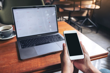 Hands holding and using mockup mobile phone with blank black screen and laptop on wooden table in cafe