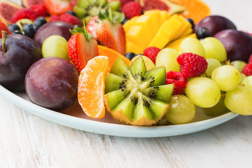 Healthy raw fruits and berries plate, strawberries raspberries oranges plums apples kiwis grapes blueberries, mango on the white woorden table, selective focus