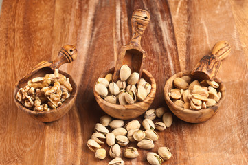Top view, medium distance of three, rare wood bowls filled with freshly roasted pistachio, almond and walnuts with artisan wood scoops on a wood serving board