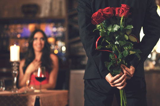 Romantic surprise concept - a man holding a bouquet of roses and wants to give it to a woman during dinner at a restaurant.