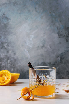 Glass of Scotch Whiskey orange juice alcohol cocktail with swirled orange peel on skewer and smoking cinnamon sticks standing on white marble table with golden holiday Christmas stars confetti.