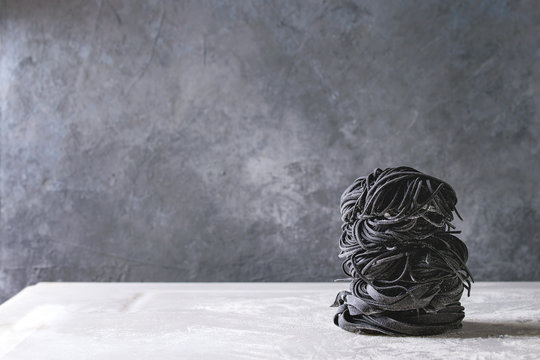 Variety Of Italian Homemade Raw Uncooked Cuttlefish Ink Black Pasta Spaghetti And Tagliatelle In Stack With Semolina Flour On White Marble Table. Copy Space.