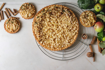 Homemade sweet apple shortbread tart on cooling rack and tartlets with cinnamon sticks, walnuts, apples branches above on white marble background. Autumn baking. Flat lay, space