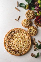 Fototapeta na wymiar Homemade sweet apple shortbread tart and tartlets with cinnamon sticks, walnuts, apples branches above over white marble background. Autumn baking. Flat lay, space