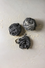 Variety of italian homemade raw uncooked cuttlefish ink black pasta spaghetti and tagliatelle with semolina flour over white marble background. Flat lay, space.