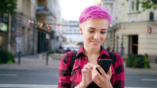 A young woman with a pink short hair using a phone in the city street. Close-up shot. Soft focus