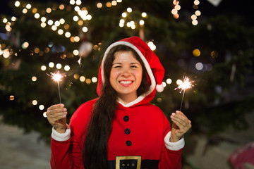 Holidays, happiness, people concept - young woman in christmas suit smiling near the christmas tree with lights in her hands