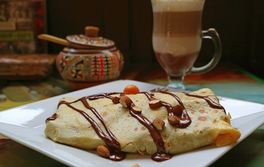 A Plate of Crepe with Chocolate Sauce and Almond Nuts, with Blurred Hot Cappuccino Coffee and Sugar Pot in Background