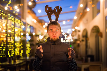 Christmas, people and winter holidays concept - surprised man in horns of deer standing at night street with bengal lights in his hands