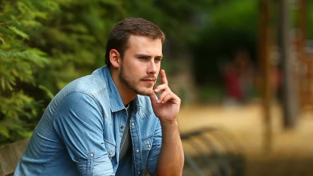 Portrait of a serious pensive guy looking away sitting on a bench in a park