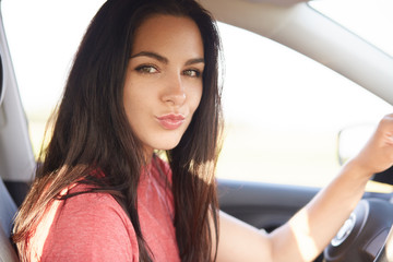 Close up shot of beautiful brunette girl pouts lips, has makeup, looks directly at camera, poses in automobile, keeps hands on wheel, makes grimace, flirts with boyfriend. Photo of female driver