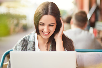 Photo of cheerful satisfied Caucasian woman looks positively at screen of laptop computer, has coverlet, reads funny stories on website, poses in outdoor cafe. People, leisure, rest concept.