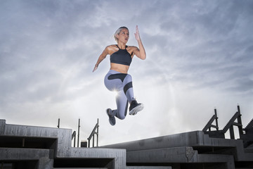Plakat Middle Eastern Girl with short braided hair jumping of a stack of blocks on a construction site wearing gray and black fitness outfit on a hot bright sunny day. 
