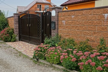 brown metal gate and brick fence near decorative green plants and flowers