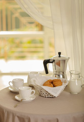 Coffee maker, cups and croissants in the basket are on a round table in front of the window.