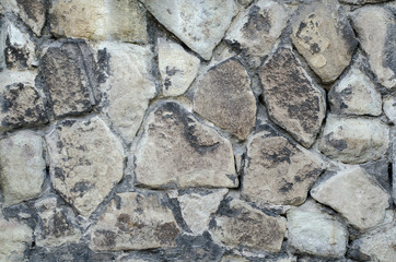 Massive light grey a stone wall with darker dirty spots. In one plane different as form but similar as dimensions stones. Old stone wall with darkened by time stones.