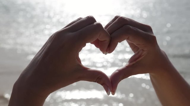 Close up view of hands of woman making heart shape at summer beach on sunrise time. Valentine day, happiness and love concept. Real time full hd video footage.