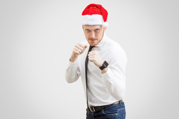 Portrait of aggressive handsome businessman in white shirt, black tie and red new year hat, looking at camera with boxing fist and serious face. indoor studio shot, isolated on light gray background.