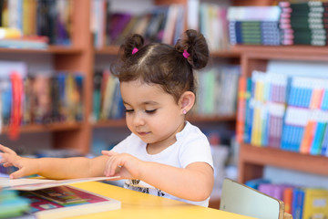 Little Girl Indoors In Front Of Books. Cute Young Toddler Sitting On A Chair Near Table and Reading Book. Child reads in a bookstore, surrounded by colorful books. Library, Shop, Shelving In Home.