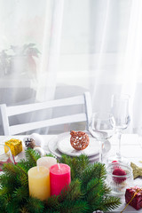 Christmas wreath with colorful big candles on a table with a Christmas table setting on a white wooden table in front of a light window
