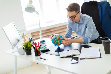 A young man sits in the office at a computer desk and holds a globe in his hands.