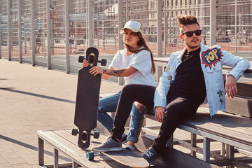 Fototapeta premium Trendy dressed young couple with skateboards sitting on a bench at city sports complex on sunny day, with warm toned.