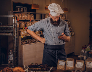 Handsome bearded chef in uniform using smartphone in kitchen.
