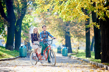 Obraz premium Young attractive tourist couple in casual clothing, bearded man and blond woman cycling on modern tandem bicycle on lit by bright sun empty pavement in beautiful autumn park under tall trees.