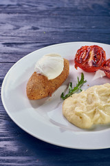 A full-fledged hot dinner on a white plate in a cafe, restaurant. Chicken Kiev stuffed with breaded, poached eggs, grilled tomato, arugula and mashed potatoes for dinner.