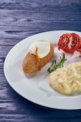 A full-fledged hot dinner on a white plate in a cafe, restaurant. Chicken Kiev stuffed with breaded, poached eggs, grilled tomato, arugula and mashed potatoes for dinner.