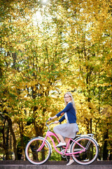 Side view of active blond long-haired attractive girl in glasses, skirt and blouse riding modern pink lady bicycle on lit by autumn sun park stairs on bright colorful golden bokeh trees background.