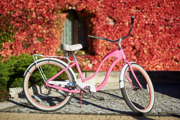 Fototapeta na wymiar Modern comfortable pink lady bicycle on sunny pavement on background of green lawn and house wall with small window fully overgrown with bright red ivy leaves. Transport and traveling concept.