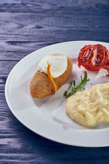 A full-fledged hot dinner on a white plate in a cafe, restaurant. Chicken Kiev stuffed with breaded, poached eggs, grilled tomato, arugula and mashed potatoes for dinner.