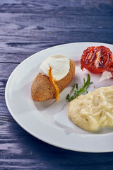 A full-fledged hot dinner on a white plate in a cafe, restaurant. Chicken Kiev stuffed with breaded, poached eggs, grilled tomato, arugula and mashed potatoes for dinner.