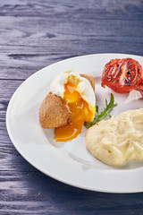 A full-fledged hot dinner on a white plate in a cafe, restaurant. Chicken Kiev stuffed with breaded, poached eggs, grilled tomato, arugula and mashed potatoes for dinner.