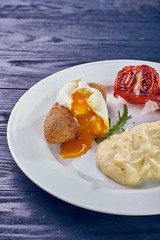 A full-fledged hot dinner on a white plate in a cafe, restaurant. Chicken Kiev stuffed with breaded, poached eggs, grilled tomato, arugula and mashed potatoes for dinner.