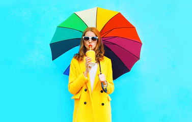 Fashion woman drinks fruit juice, holds colorful umbrella in yellow coat jacket on blue background