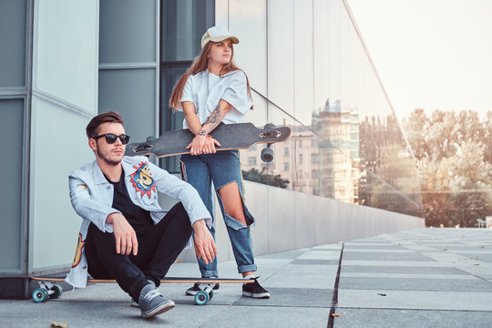 Trendy dressed couple - young hipster man sitting on a longboard and his girlfriend standing near outdoors.