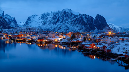 Lofoten islands, Norway. Colorful winter landscape in blue hours. Illuminated fishing village reflected in water. Snowy mountains in background. 