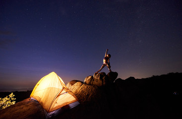 Camping site in mountains. Bright tourist tent on steep rock and young slim woman posing with raised arm on dark blue starry evening sky. Tourism, yoga, active lifestyle, climbing concept.