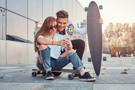 Trendy dressed couple with skateboards views interesting photos together on smartphone on the street.