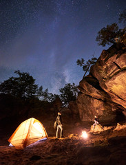 Camping night in valley amid huge rock formations. Young tourist couple, standing near tent. Young woman sitting on big boulder looking at bonfire under dark starry sky. Tourism and hiking concept.