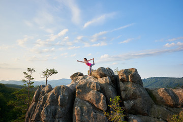 Young slim woman doing complicated yoga pose on lit by summer sun top of huge rock on green tree tops and clear blue sky background in the morning. Fitness and healthy lifestyle concept. Narajasana