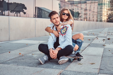 Trendy dressed young couple - pretty girl embrace her boyfriend while they sitting together on a skateboard near skyscraper.